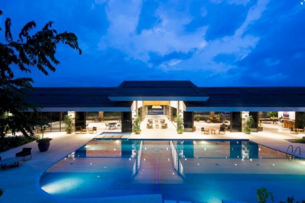 A luxurious resort pool area at dusk, featuring a large pool, lounge chairs, and an open-air building with lights.