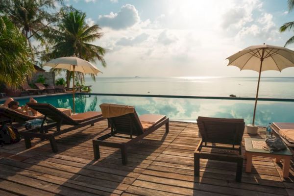 Deck with sun loungers and umbrellas by a pool, overlooking the ocean, with palm trees and a bright sky in the background.