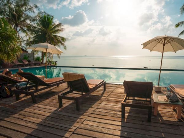 Deck with sun loungers and umbrellas by a pool, overlooking the ocean, with palm trees and a bright sky in the background.