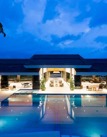 The image shows a luxurious building with an illuminated pool at night, surrounded by lounge chairs and greenery, under a partly cloudy sky.