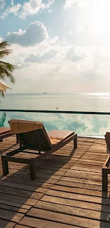 Lounge chairs and umbrellas by an infinity pool, overlooking a scenic ocean view with palm trees.