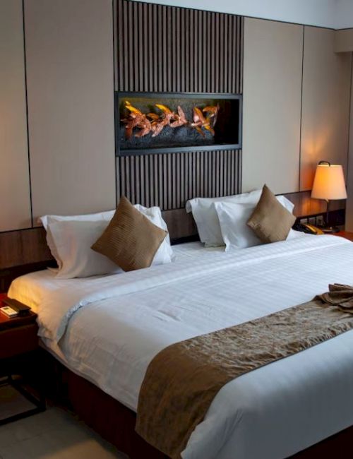 A cozy hotel room features a large bed with white linens, bedside lamps, and a decorative wall piece. A window is covered with curtains.