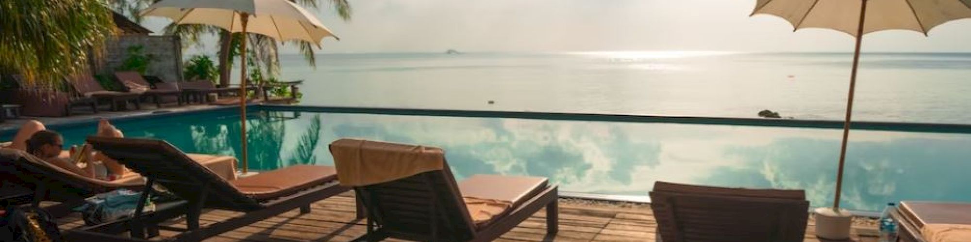 A serene poolside scene with sun loungers, umbrellas, palm trees, and a view of the ocean under a sunny sky.