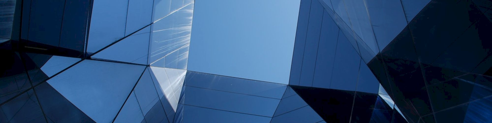 The image shows a geometric arrangement of reflective glass panels forming a structure with a clear blue sky visible in the center.