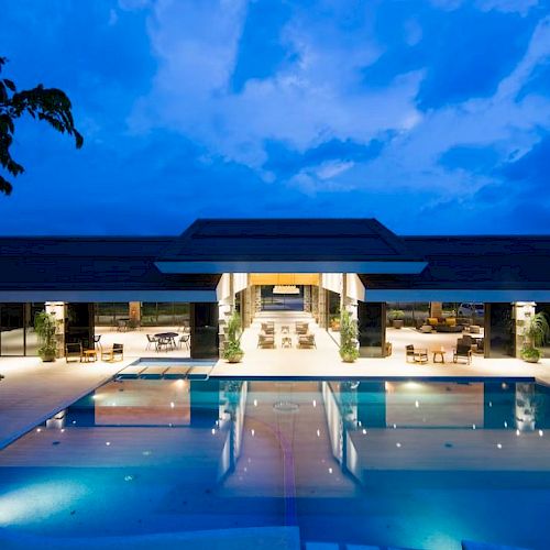 The image shows a luxurious, well-lit villa with an outdoor pool and calm evening ambiance.