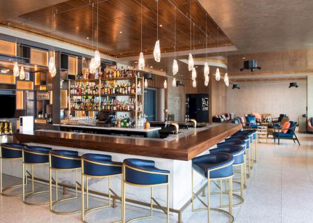 A modern bar with blue chairs, a well-stocked bar counter, hanging lights, and seating area in a spacious, stylish setting.