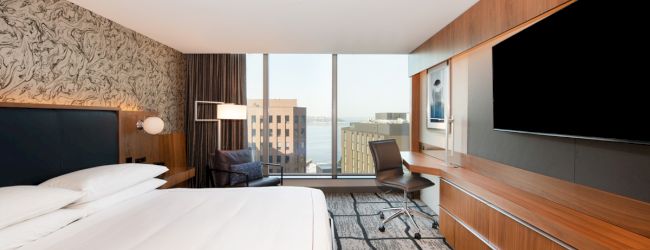 A modern hotel room with a large bed, desk, chair, TV, and a window offering a city view. The decor is sleek with wood accents and patterned walls.