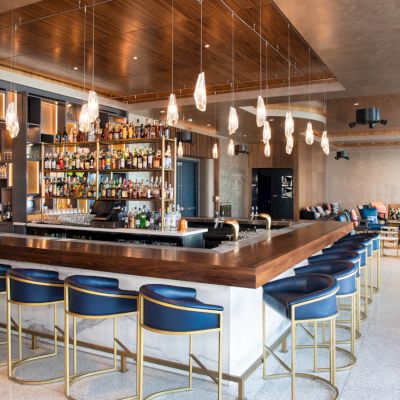 A modern bar with blue stools, pendant lights, and a well-stocked liquor shelf, set in a spacious lounge area with cozy seating.