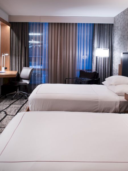 The image shows a modern hotel room with two beds, a wall-mounted TV, a desk with a chair, and decorative lighting on a patterned wall.