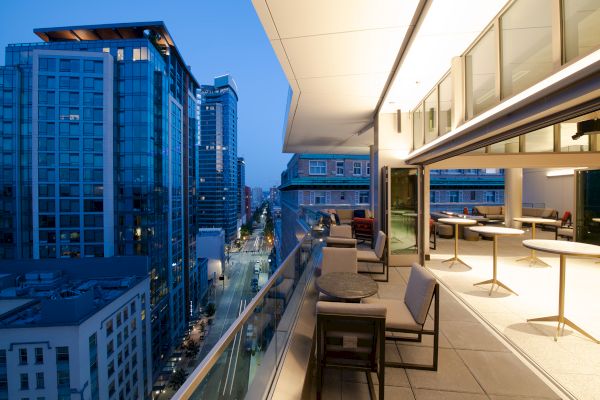 A modern cityscape with tall buildings and a sleek rooftop terrace featuring seating and tables, overlooking a city street at dusk.