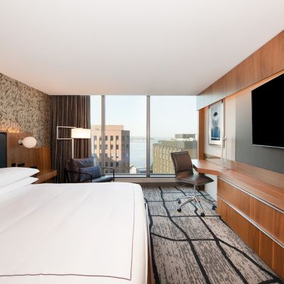 A modern hotel room with a large bed, wall-mounted TV, desk, and city view through the window, featuring stylish decor and a patterned carpet.