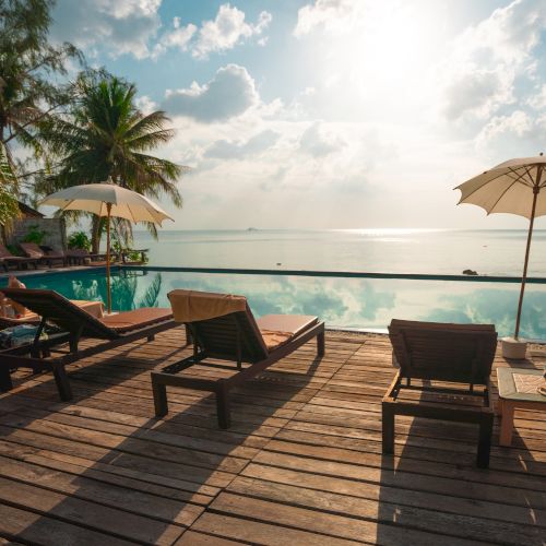 A serene poolside scene with lounge chairs, umbrellas, and palm trees overlooking a calm ocean under a bright sky.