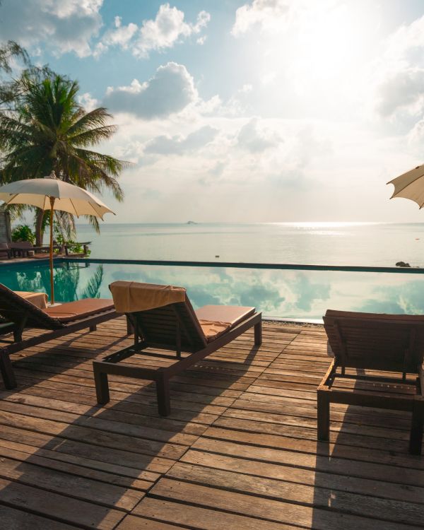 A serene poolside scene with lounge chairs, umbrellas, and palm trees overlooking a calm ocean under a bright sky.