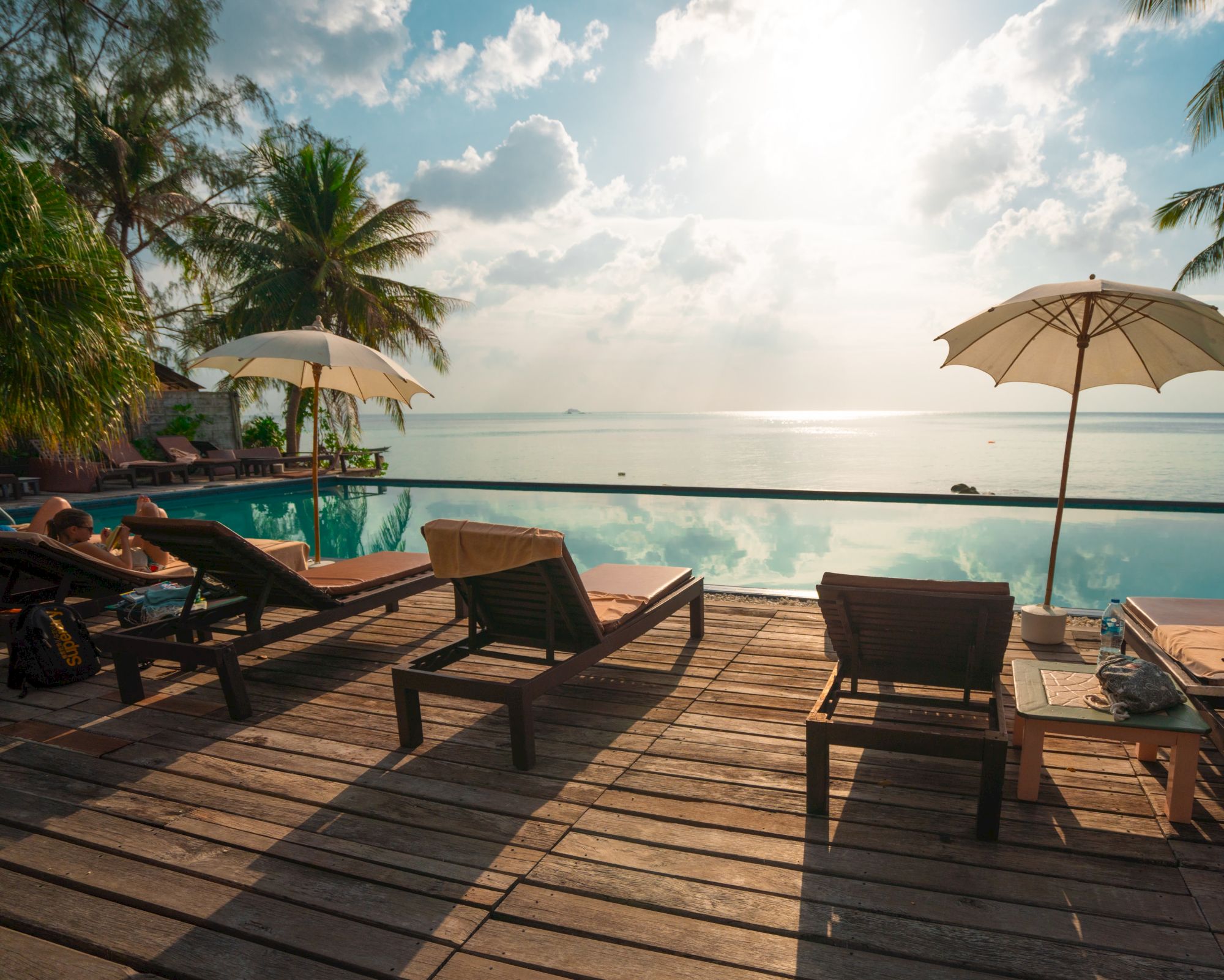A serene beach scene with lounge chairs and umbrellas by an infinity pool facing the ocean, under a partly cloudy sky and bright sun.