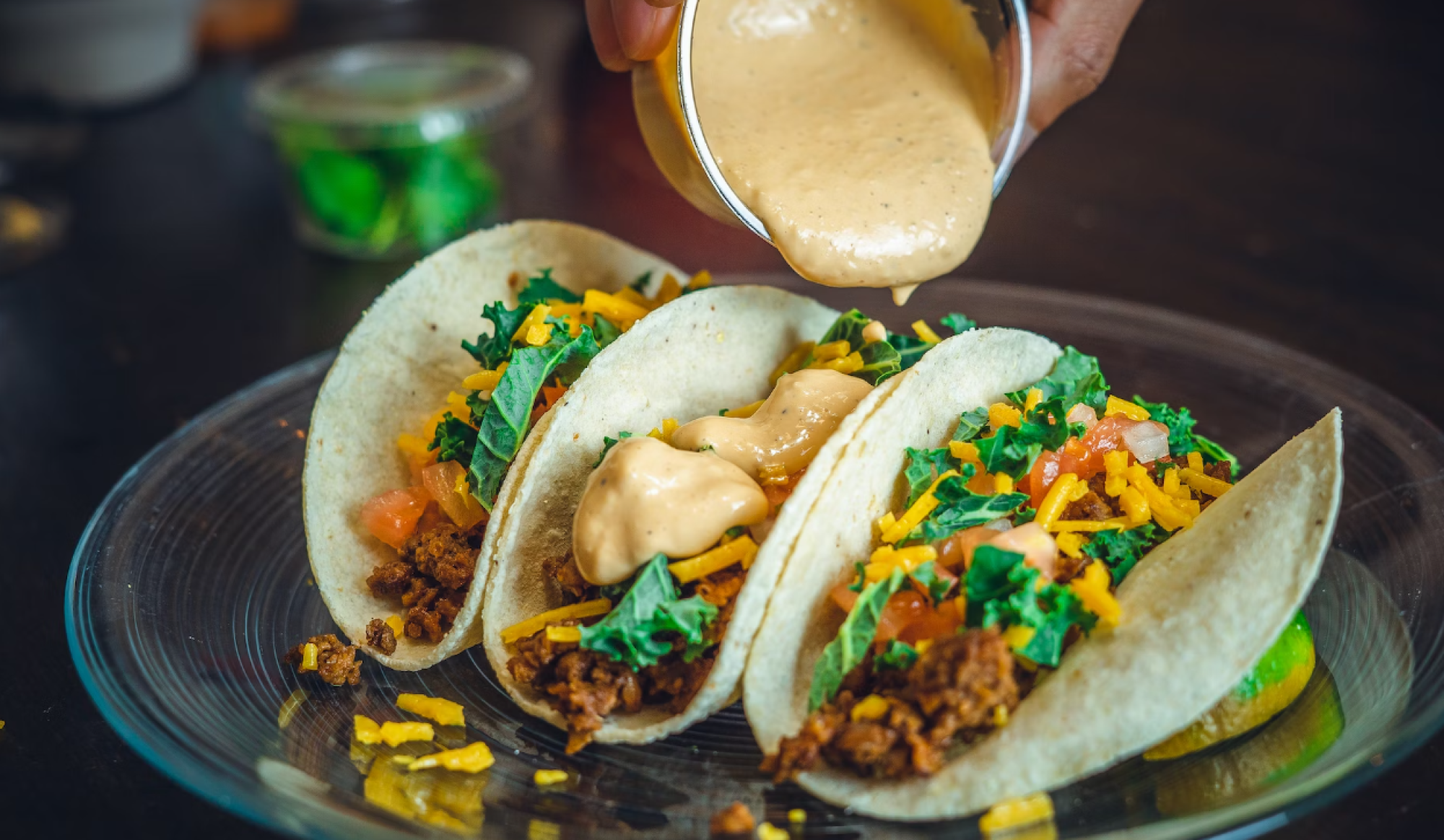 Three tacos filled with ground meat, cheese, tomato, and greens, topped with sauce being poured from a cup.