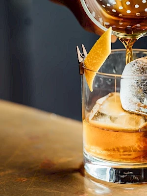 A hand pours a brown liquid into a glass with ice and a citrus garnish, using a strainer, on a bar surface.