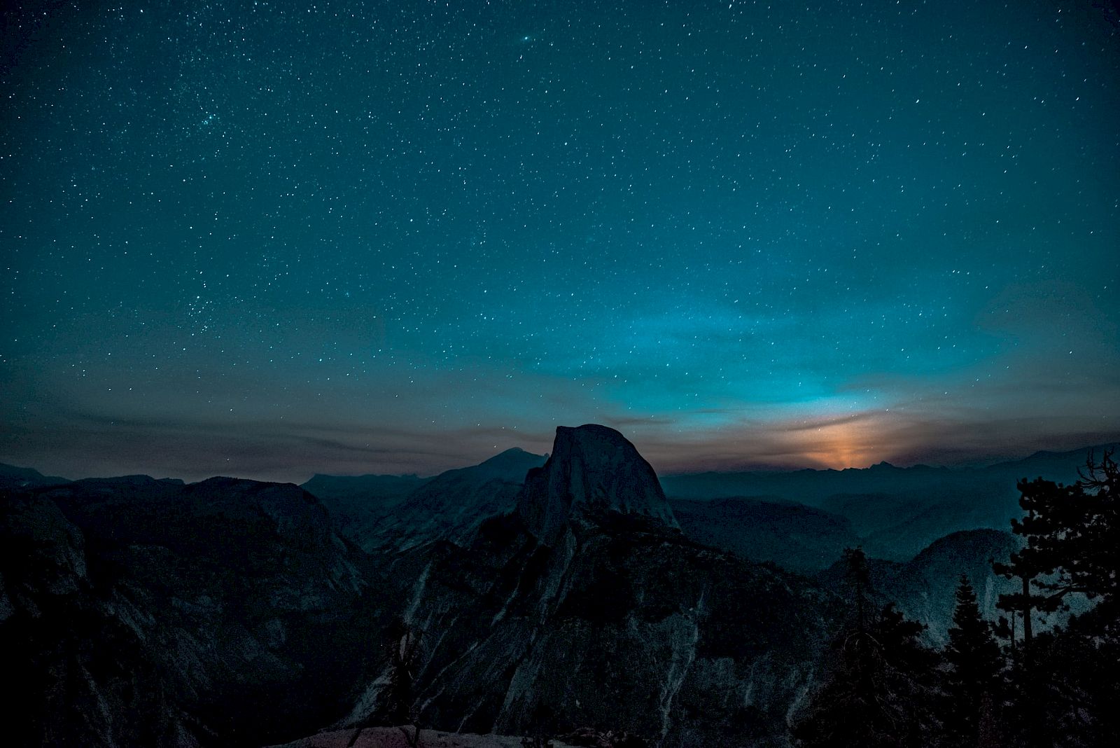 A starry night sky over a mountainous landscape with trees and a glow on the horizon, possibly from a setting or rising moon.