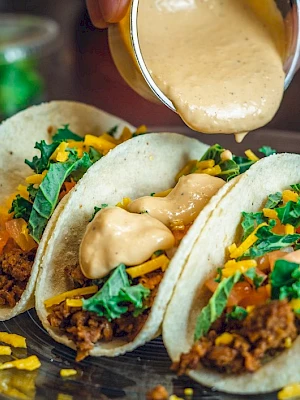 Three tacos with ground meat, cheese, lettuce, and tomatoes, being topped with a creamy sauce on a plate.