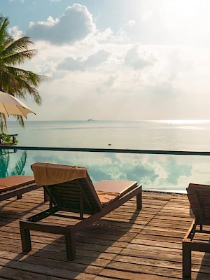 A relaxing poolside scene with lounge chairs, umbrellas, and palm trees overlooking a serene ocean view under a partly cloudy sky.