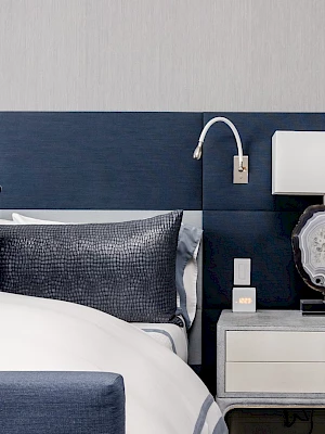 A modern bedroom with a blue headboard, white bedding, pillows, and a side table featuring a lamp and wall-mounted reading light.