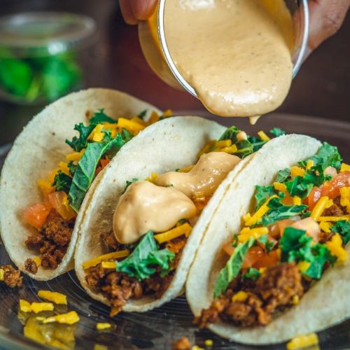 Three tacos filled with meat, cheese, tomato, lettuce, and topped with sauce being poured over them.