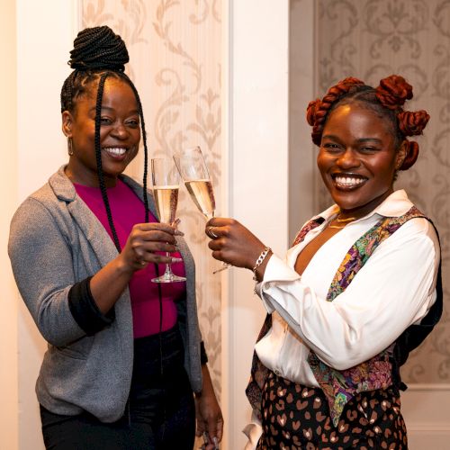Two people smiling and clinking champagne glasses in a warmly lit room with patterned wallpaper.