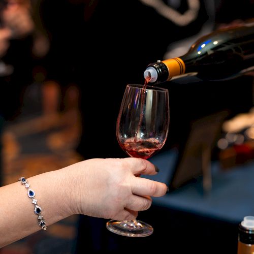 A person is pouring red wine from a bottle into a glass held by another person at an event.