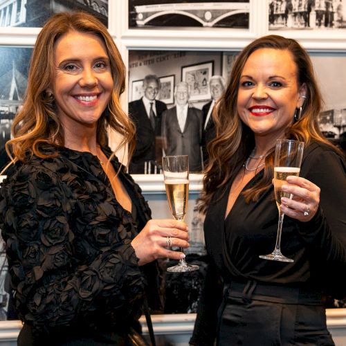 Two women in black outfits holding champagne glasses, smiling, with black-and-white photos on the wall behind them.