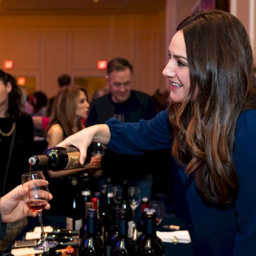 A woman is pouring wine at a social event with people in the background holding glasses and mingling.