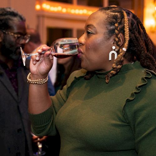 A person in a green sweater sips from a wine glass at a social gathering, with others in the background and soft lighting.