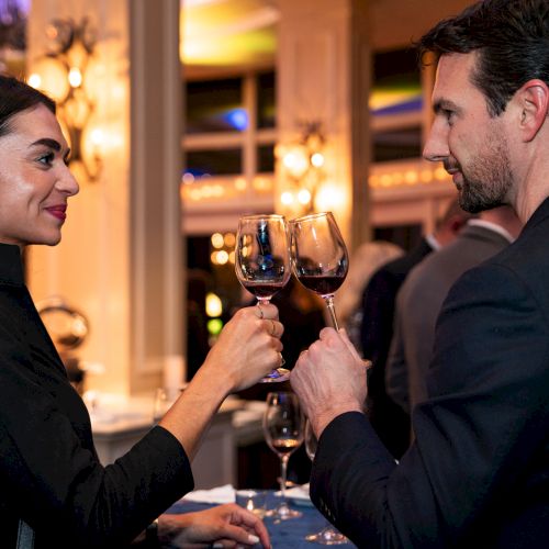 Two people are clinking wine glasses together in a warmly lit, elegant setting.
