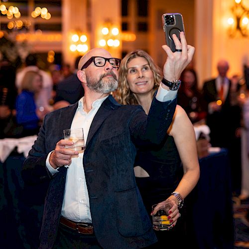 A man and woman take a selfie at an elegant event, holding drinks and posing with a crowd in the background.