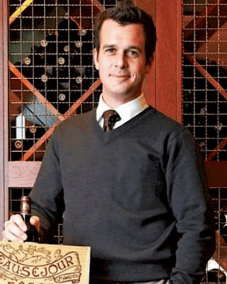 A person stands in front of a wine rack, holding a wooden wine box labeled "CHATEAU SAINT GEORGES." They are wearing a dark sweater and tie.