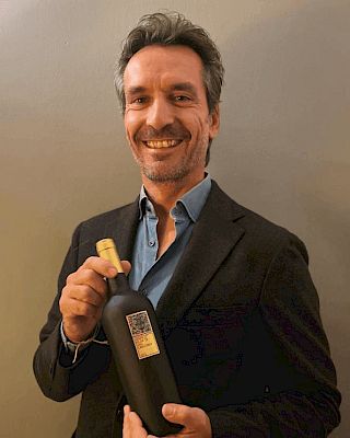 A man in a suit is smiling and holding a bottle with a label, standing against a plain background.