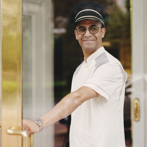 A person in a uniform, possibly a doorman or concierge, is smiling and opening a door.