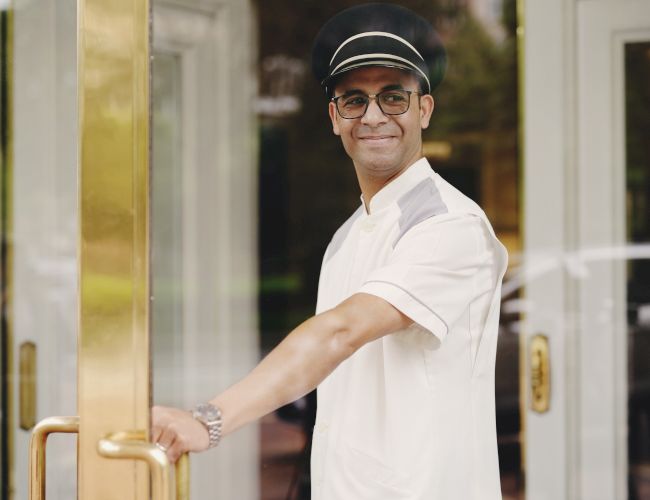 A person in a uniform, possibly a doorman or concierge, is smiling and opening a door.