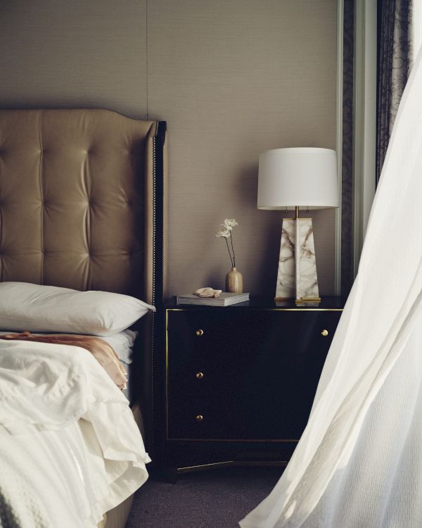 A stylish bedroom corner with a tufted bed, a nightstand, a lit table lamp, and floral decor beside patterned curtains.