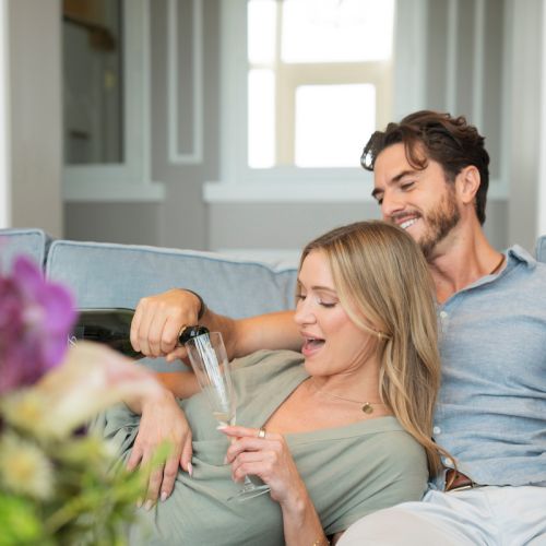 A couple relaxes on a couch at home, toasting with champagne flutes while smiling in a bright living room.