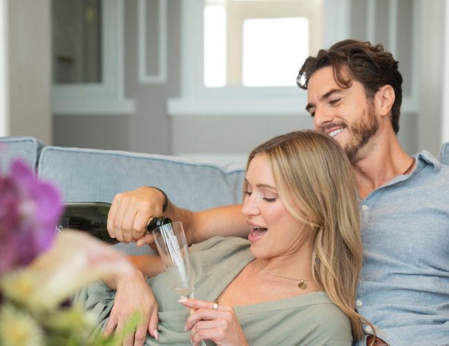 A couple relaxes on a couch at home, toasting with champagne flutes while smiling in a bright living room.