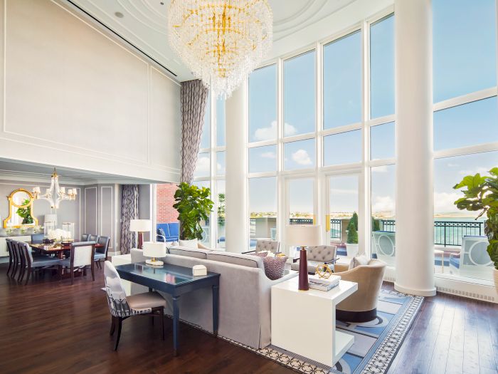 This image shows a luxurious, open-plan living and dining room with tall windows, elegant furnishings, chandeliers, and large potted plants.