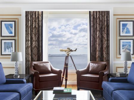 A living room with two brown chairs, a telescope by the window, blue sofas, and framed art on the walls. The view shows a body of water.