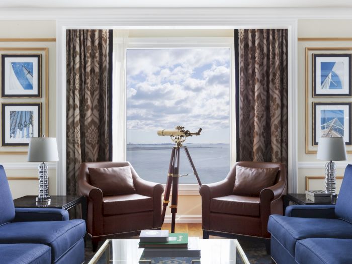 A living room with two brown chairs, a telescope by the window, blue sofas, and framed art on the walls. The view shows a body of water.