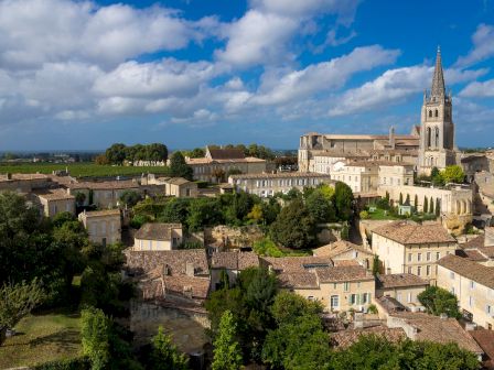 A scenic view of a historic town with stone buildings, nestled among greenery, featuring a prominent church with a tall spire under a blue sky.