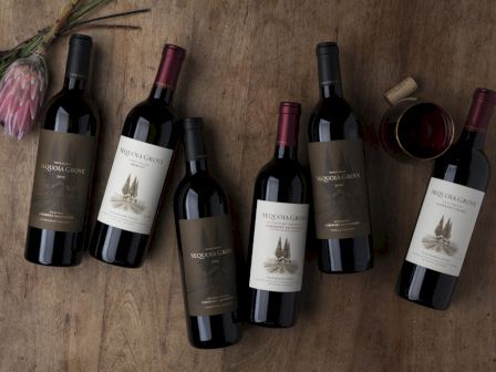 Six wine bottles on a wooden surface with a glass of red wine and flowers beside them.