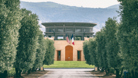 A building with a curved driveway, two flags, surrounded by trees, and hills in the background, under a cloudy sky.