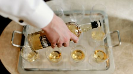 A person in a white shirt is pouring a bottle of liquor into multiple glasses on a tray.
