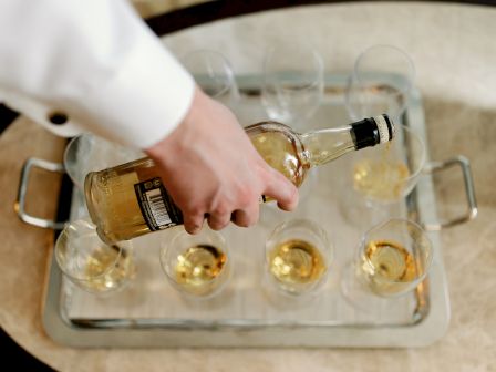 A person in a white shirt is pouring a bottle of liquor into multiple glasses on a tray.