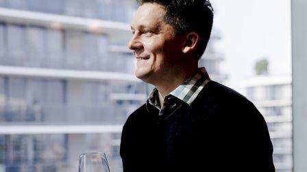 A man in a dark sweater gazes out a window with a glass of red wine, smiling contently against a blurred cityscape backdrop.