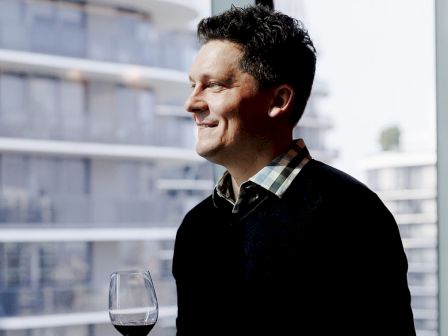 A man in a dark sweater gazes out a window with a glass of red wine, smiling contently against a blurred cityscape backdrop.