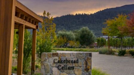 The image shows a stone sign that reads "Cakebread Cellars" with a scenic background of trees and hills under a cloudy sky.